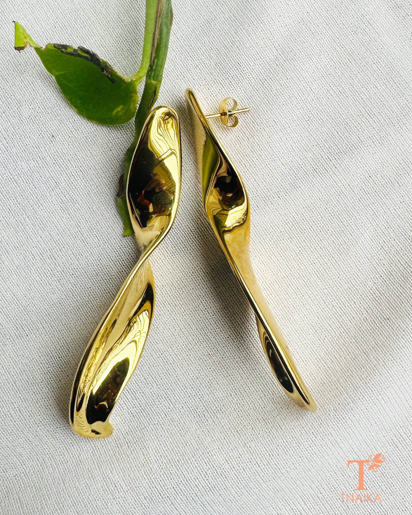 Long sculptural gold earrings styled on a white fabric background with green leaves.