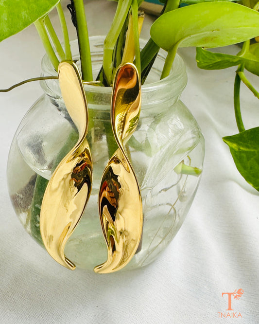 Sculpted gold-tone drop earrings with a curved fluid design displayed against a green plant.