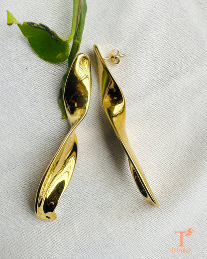 Long sculptural gold earrings styled on a white fabric background with green leaves.